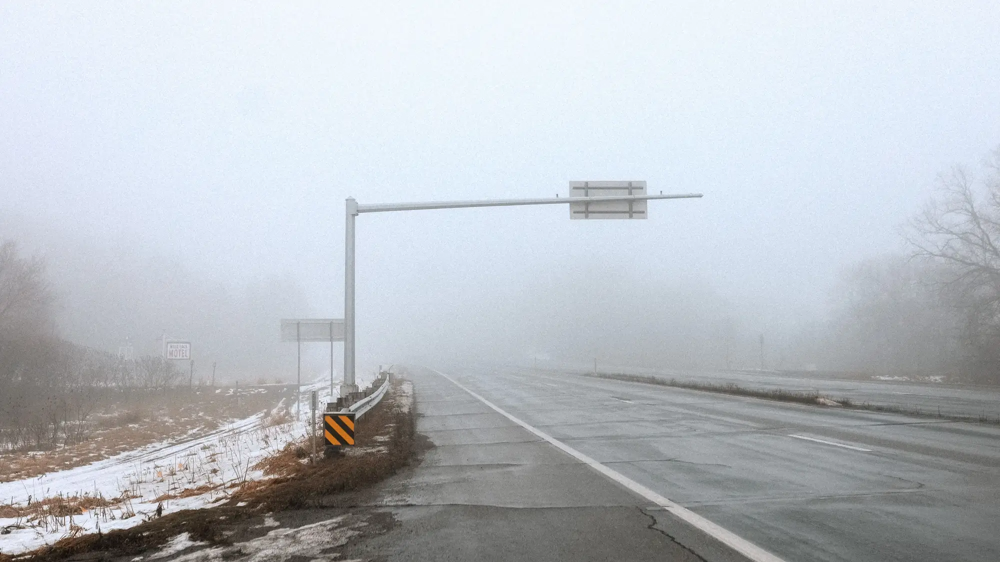 A divided highway disappearing into winter fog, an overhead sign arm at the shoulder and no count station in sight